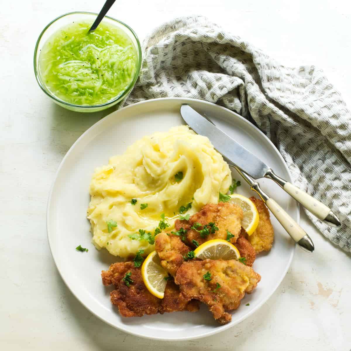 Czech schnitzel with mashed potatoes and a bowl of cucumber salad on a side.