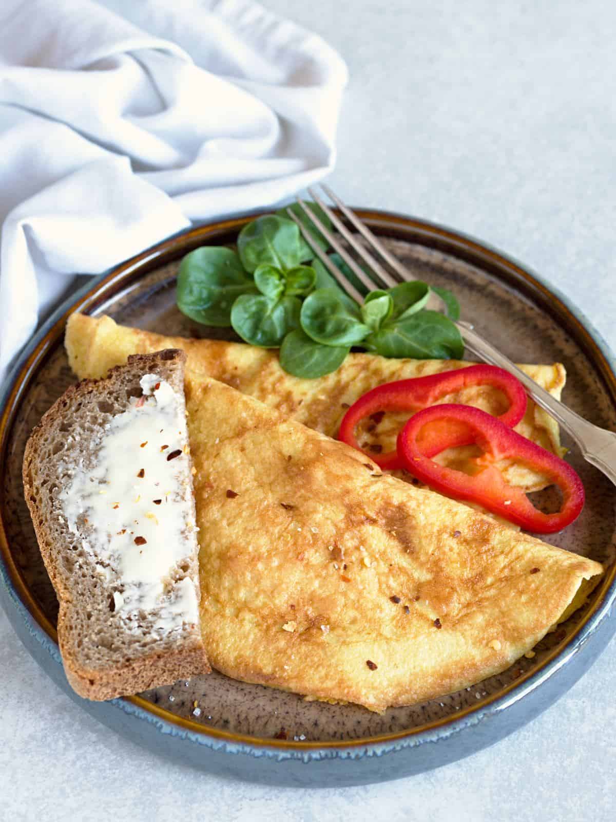 Fluffy omelette with red paprika, green leaves and a piece of buttered bread.