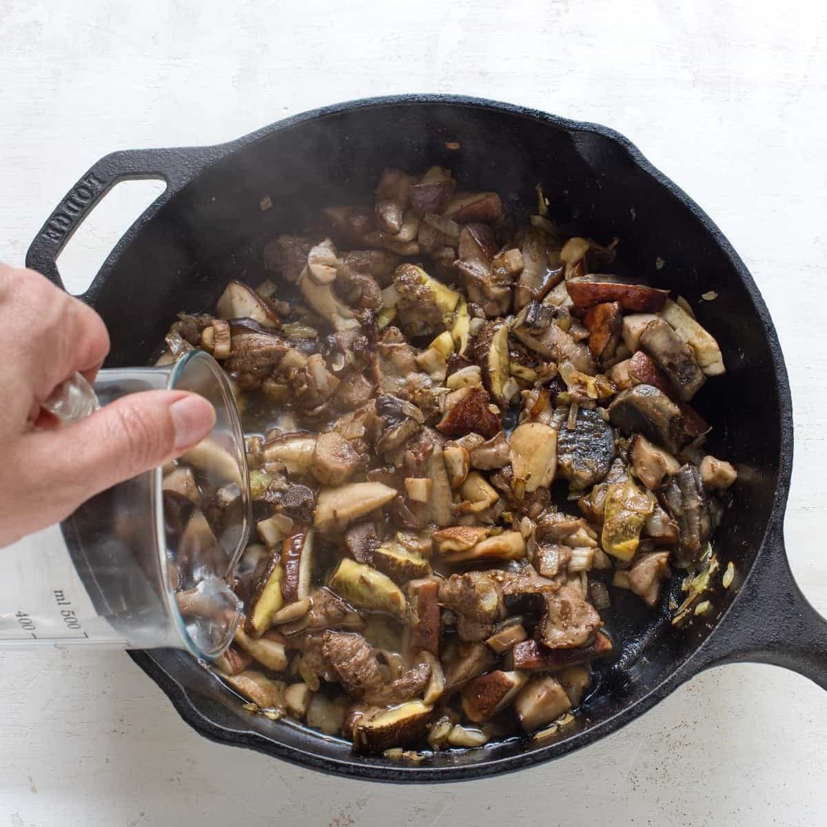 Pouring water to a skillet with sauteed mushrooms.