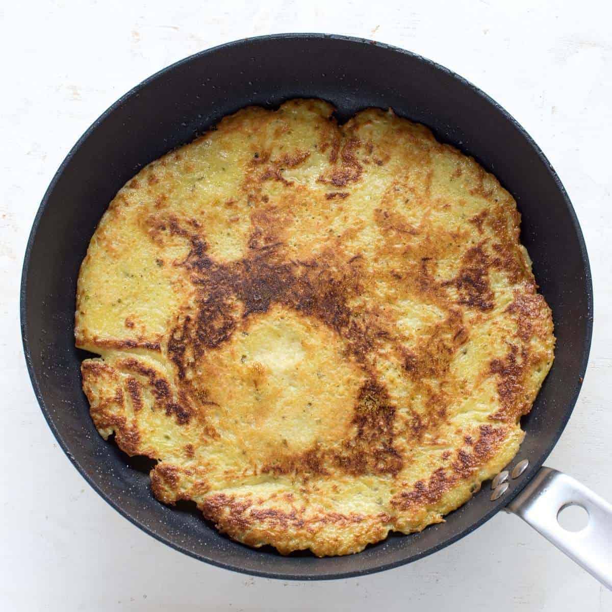 A potato pancake is frying in a pan.