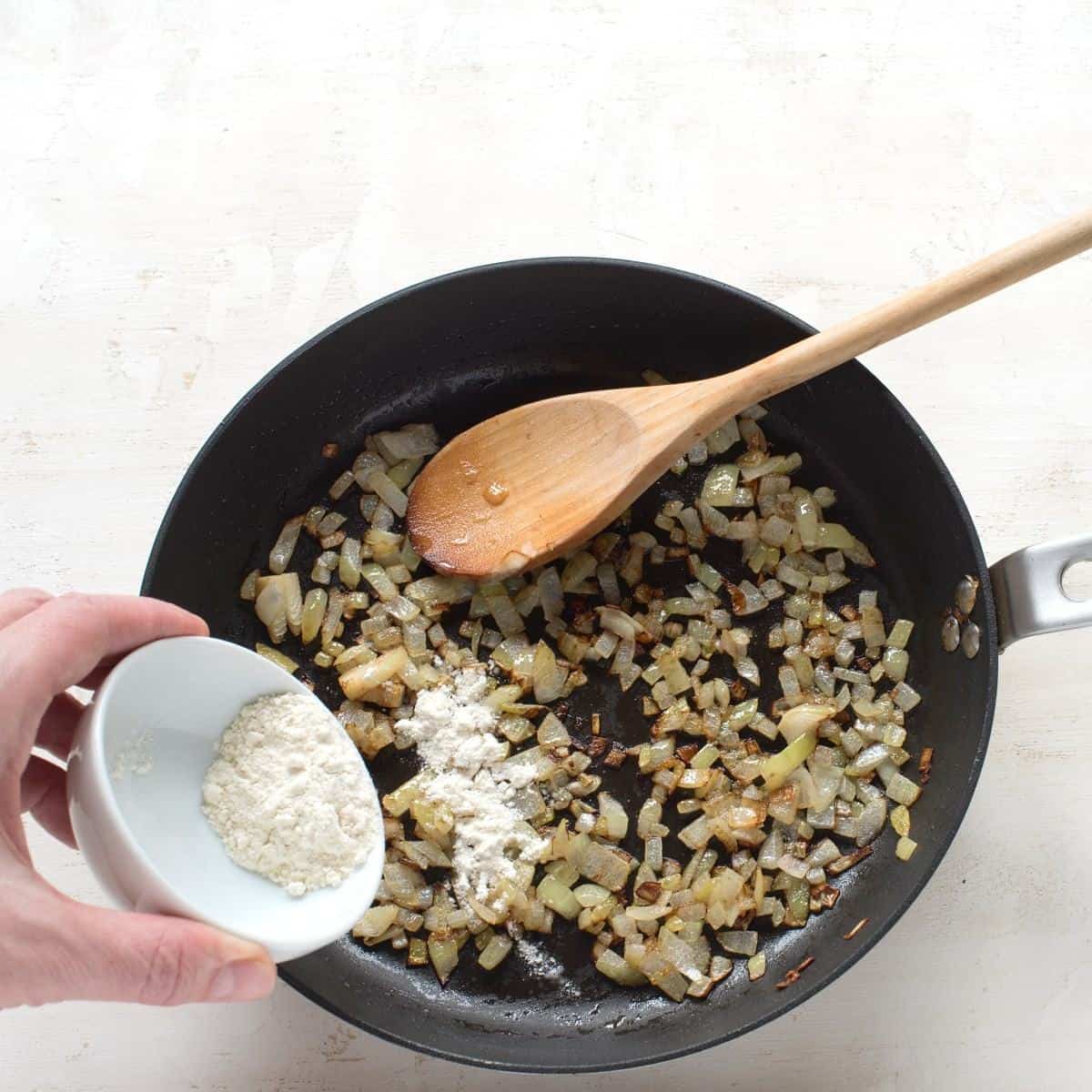 Pouring flour to fried onion in a black skillet.