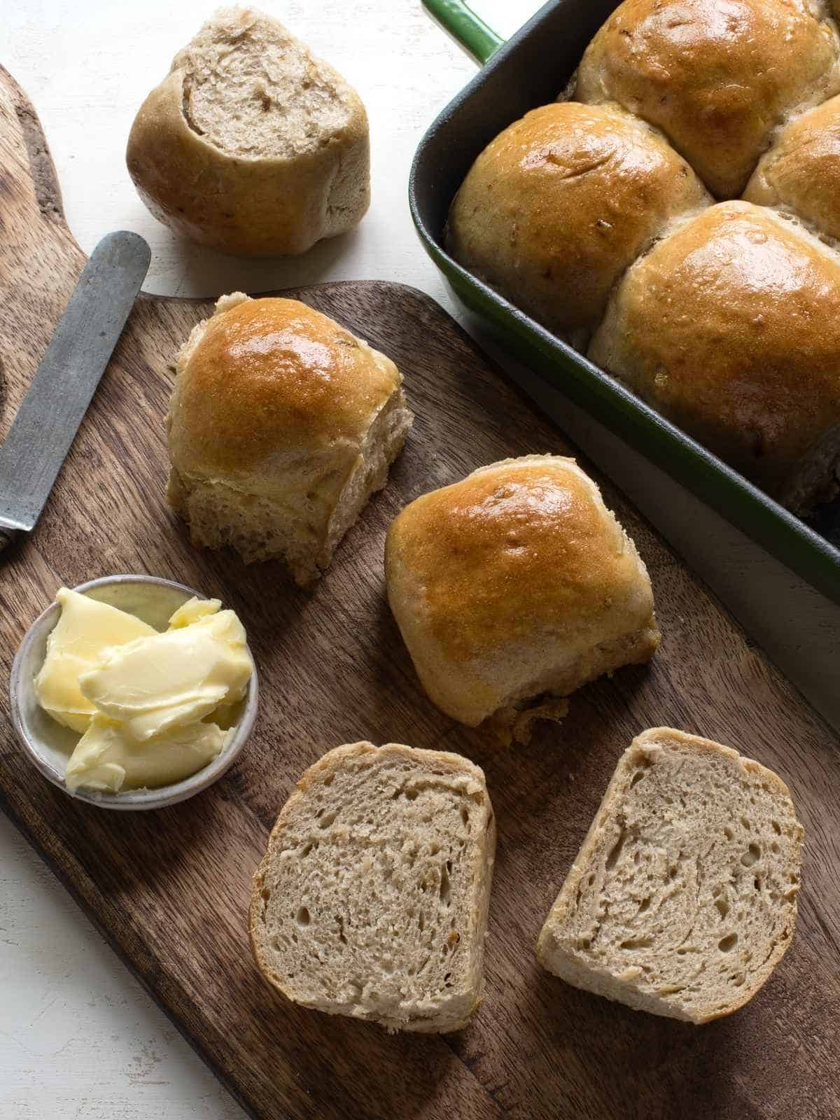 Baked onion bread rolls, one of them cut in two halfes. On a brown wooden board, with a small bowl of butter.