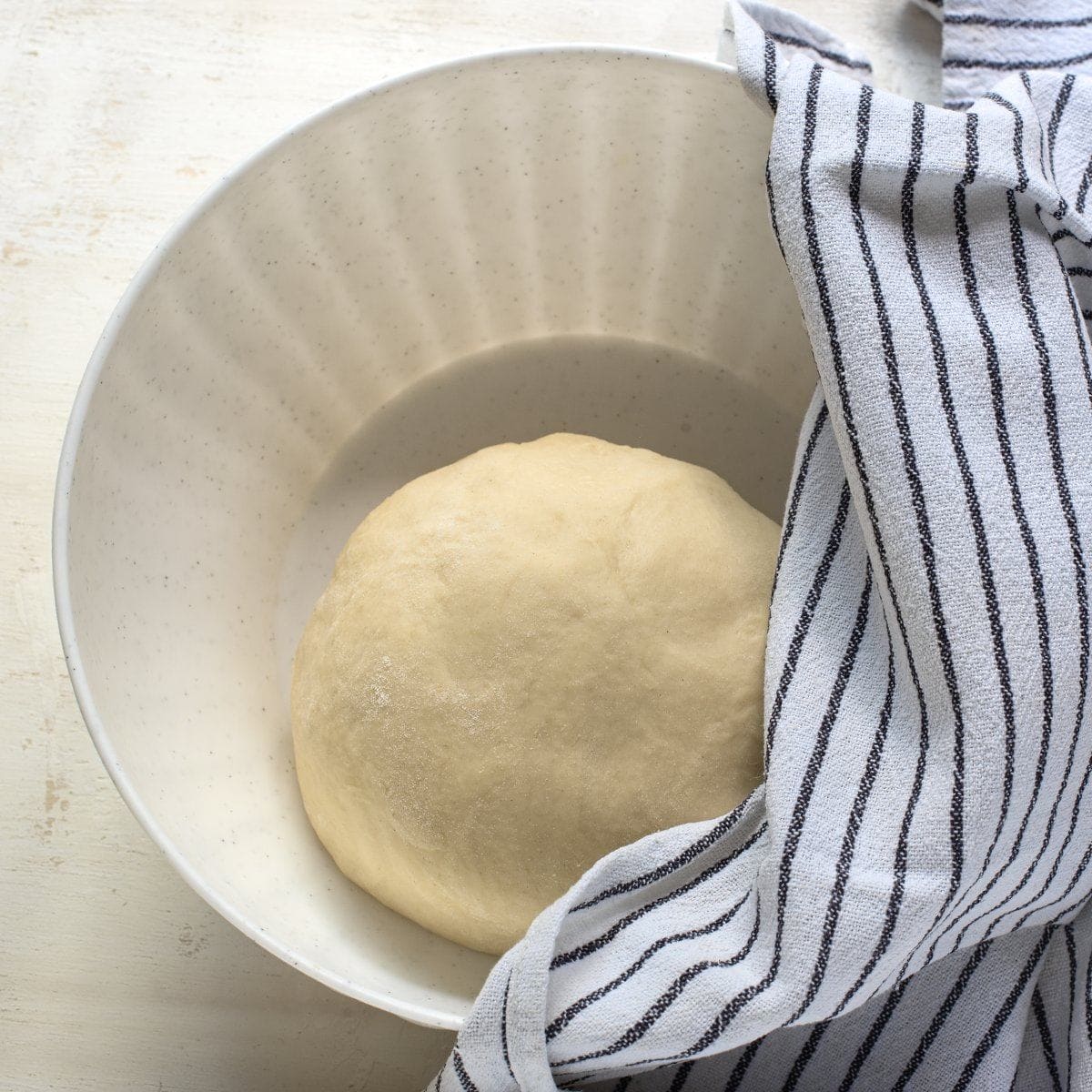Ball of yeast dough before rising. In a white bowl, partly covered with stripped kitchen towel.