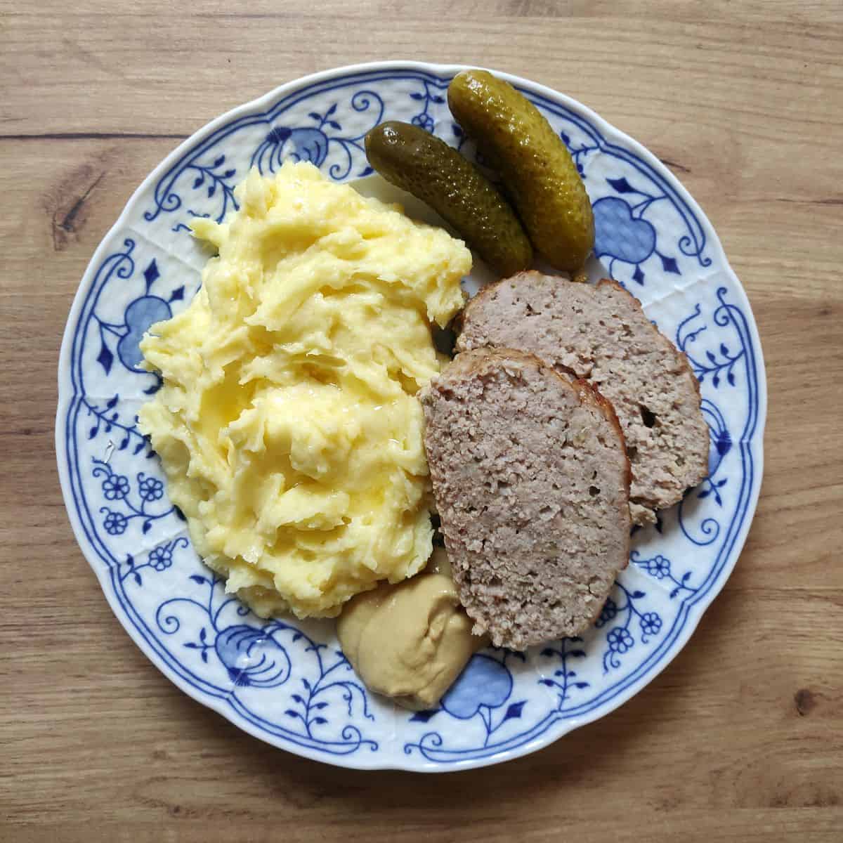 Two slices of Czech sekaná served with potato puree, whit yellow mustard and pickles as sides.