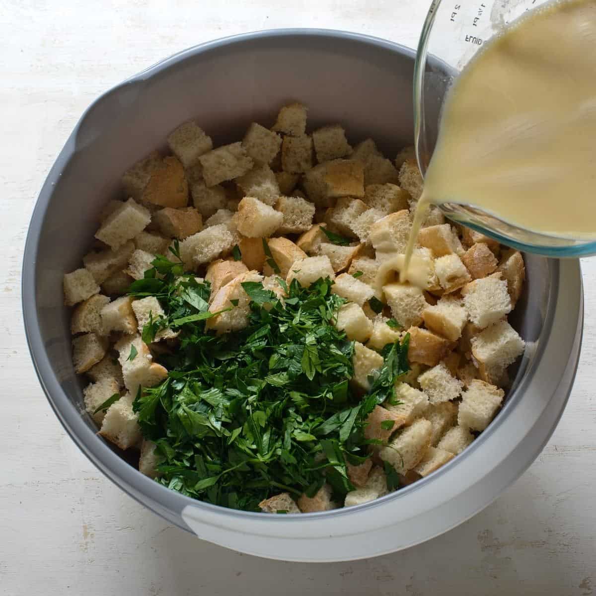 Pouring eggy mixture into a bowl of bread cubes and chopped parsley.